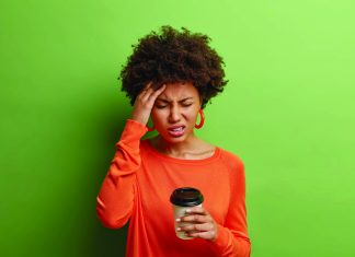 Stressful Afro American woman feels terrible headache smirks from pain closes eyes touches temple drinks coffee wears casual orange jumper isolated over vivid green background. Health problems