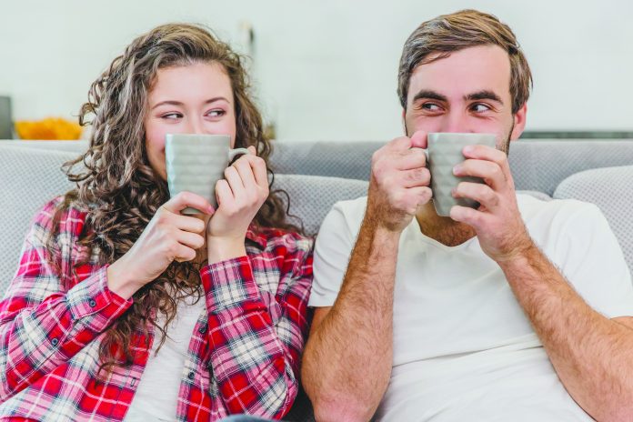 A beautiful couple of young people. Funny people raised the cup of coffee with her face closed. During this, they look at the camera and smile. Sitting on the couch at home.