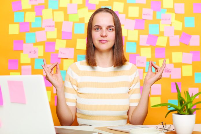 Woman sitting at table, working on laptop, standing with raised arms and doing yoga meditating exercise, practicing yoga mudras to relax and concentrate against yellow wall covered with sticky notes.