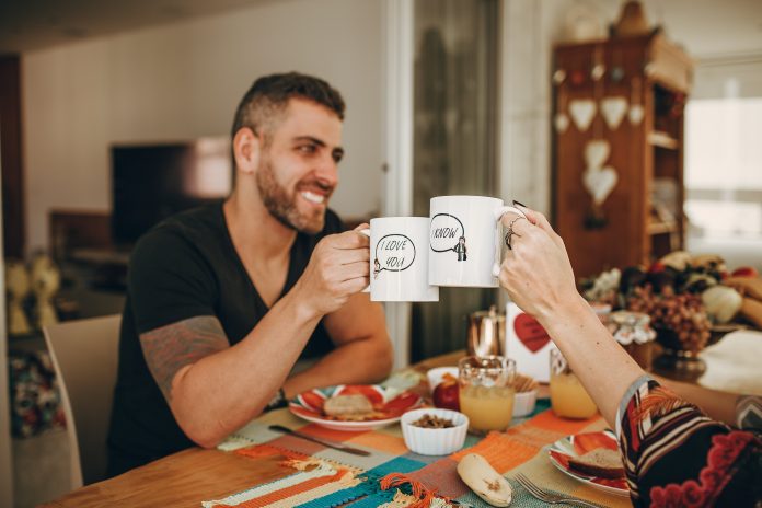 Two people happily clinking mugs across a kitchen table